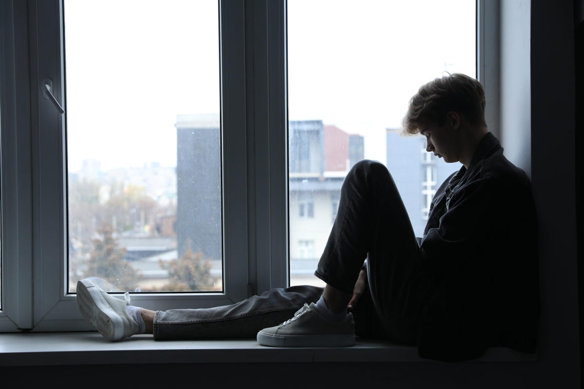 Depressed teenage boy sitting on window sill and looking down