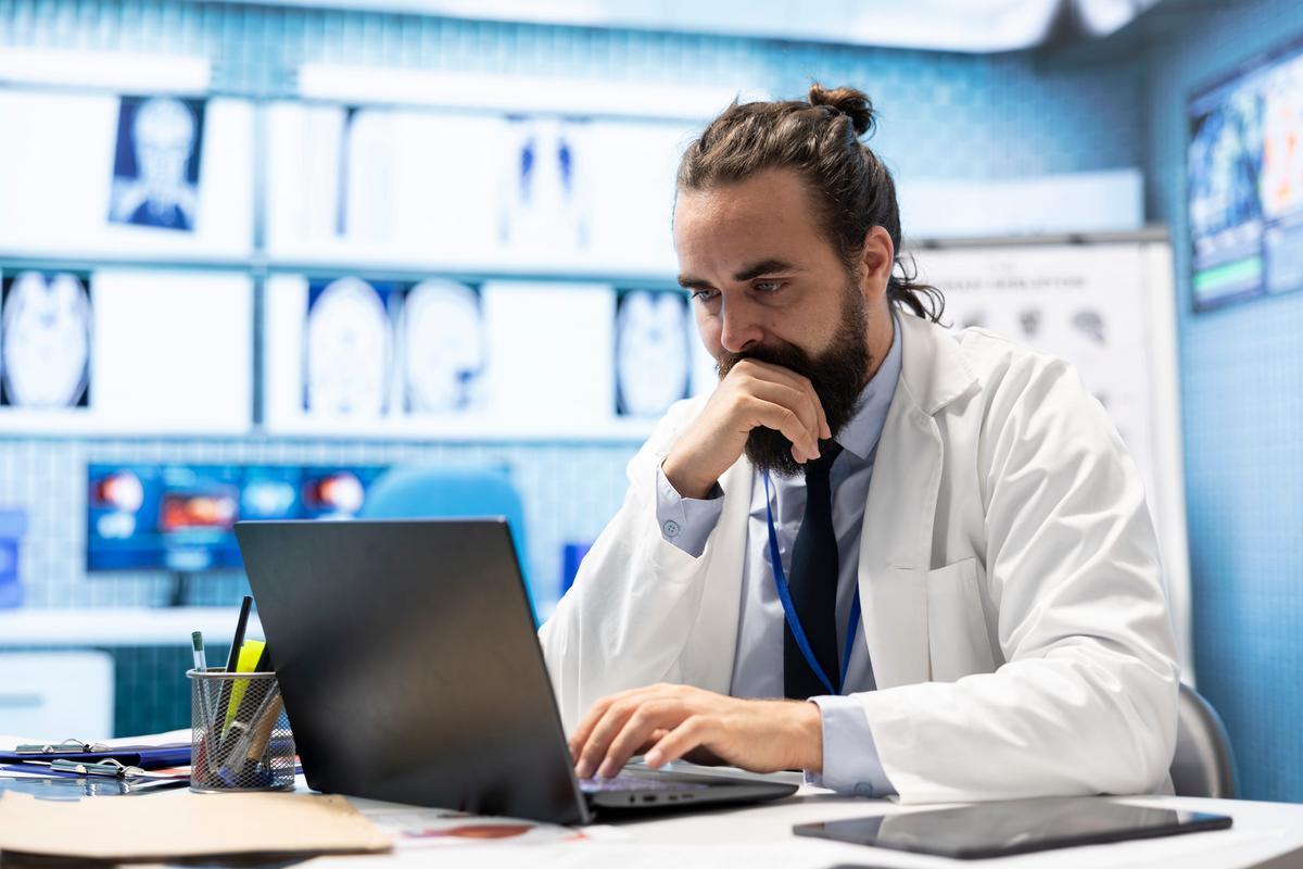 A clinician in a white coat working at a laptop in a medical office.