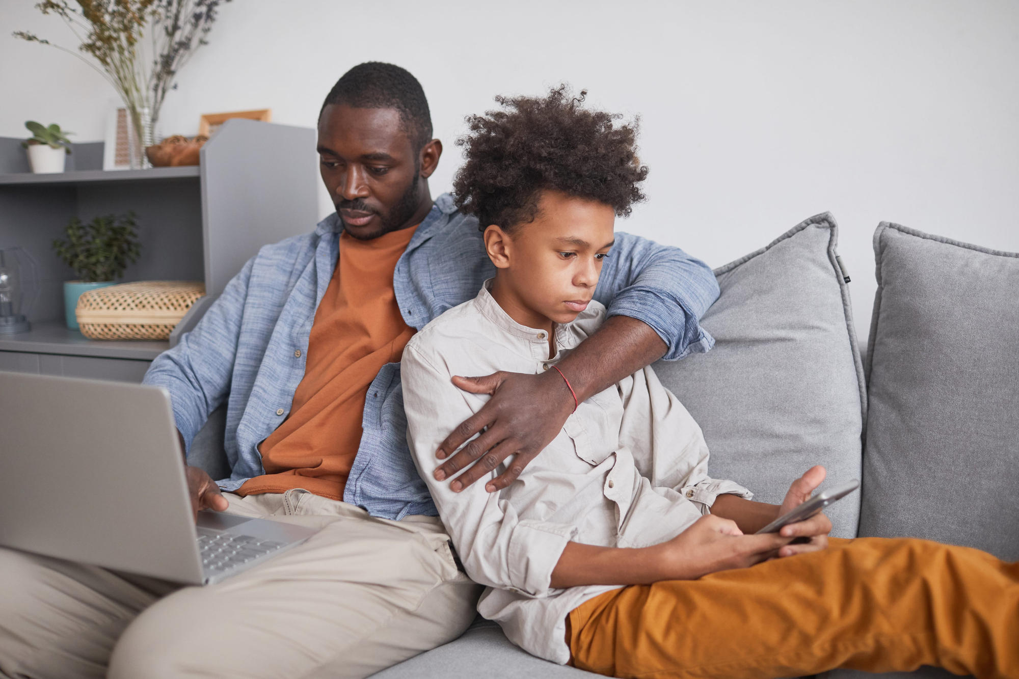 Man and his teen son sitting together on sofa in living room surfing Internet using laptop and smartphone
