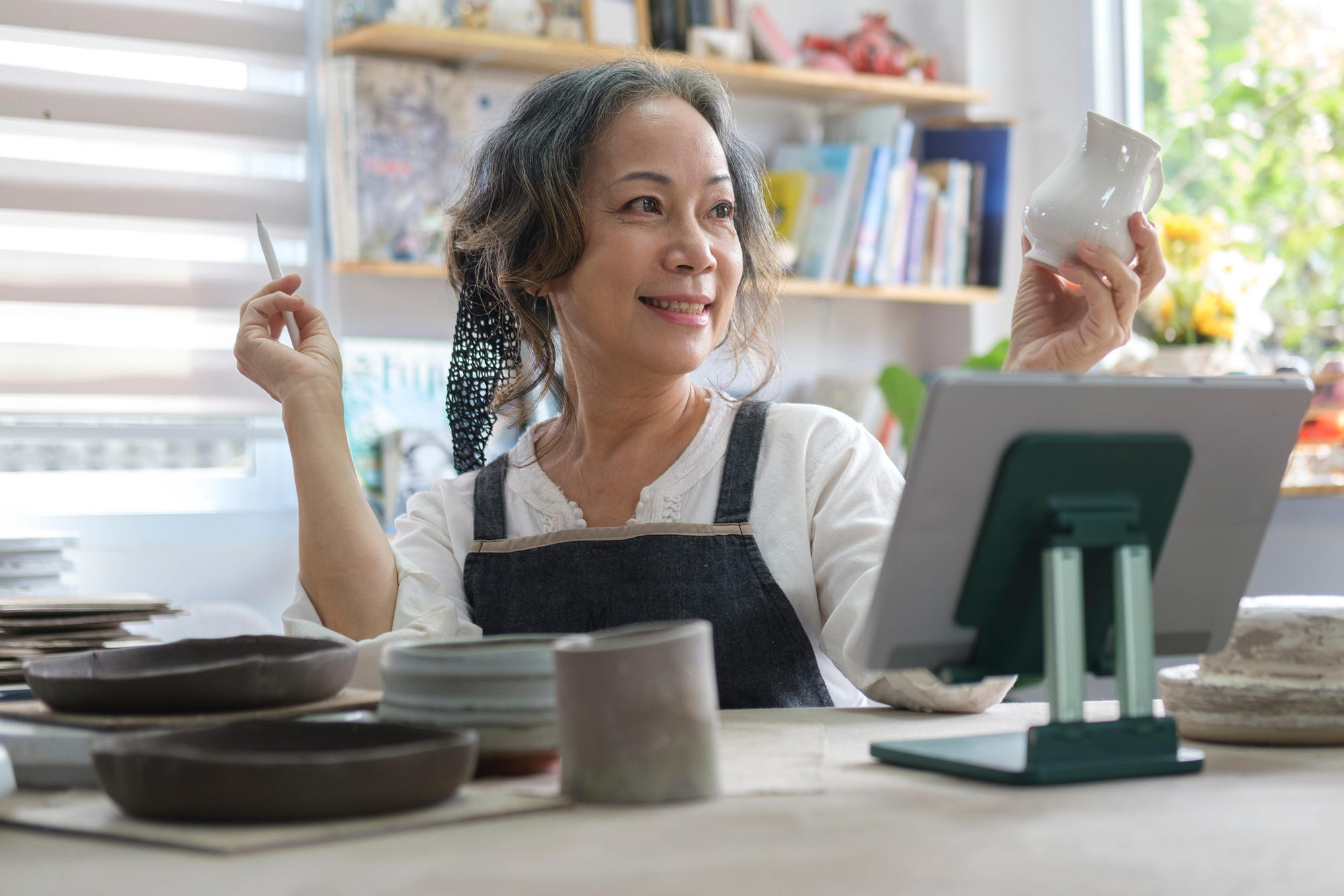 Smiling senior female holding pottery earthenware product in front of digital tablet